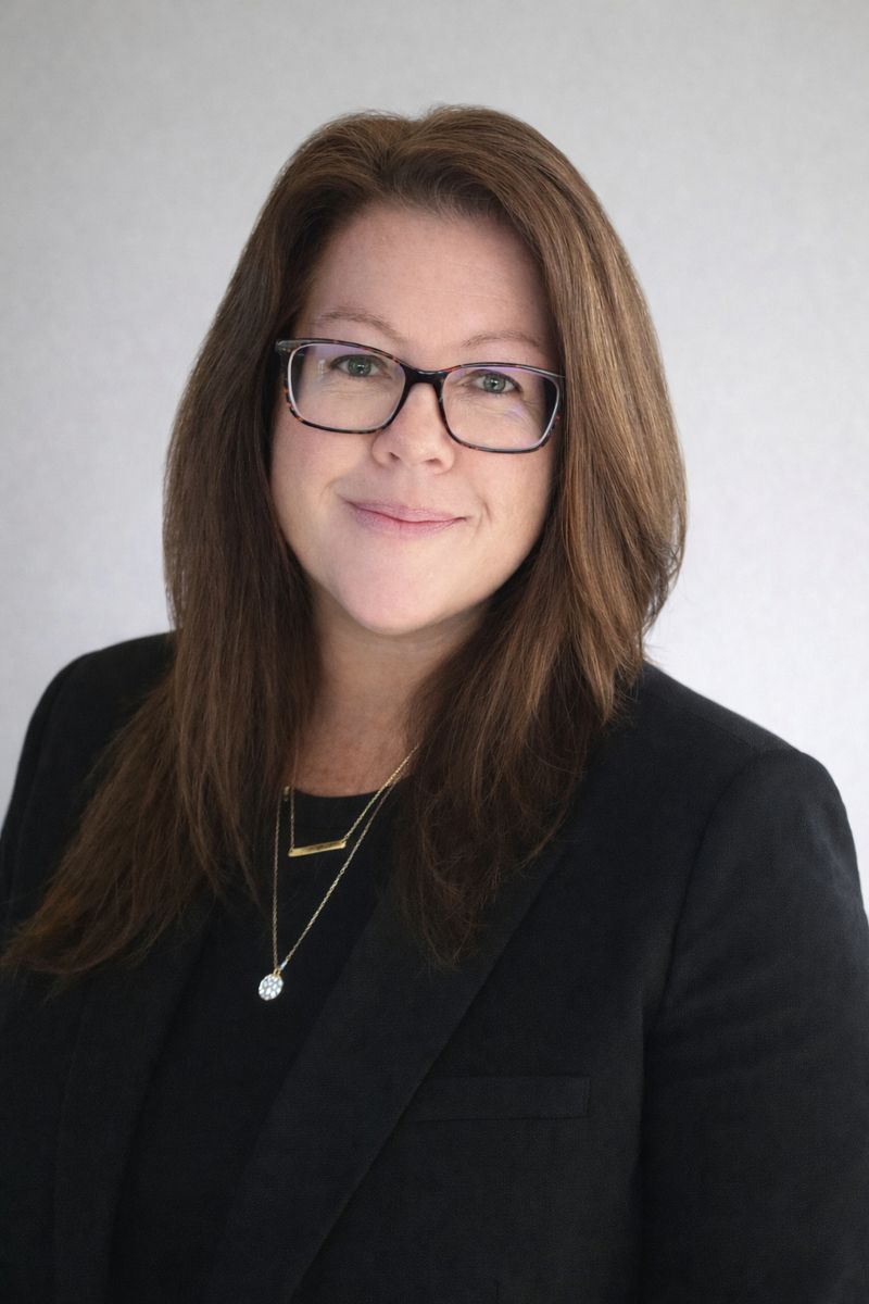 Professional headshot of a woman with long brown hair, wearing glasses and a black blazer against a neutral background.